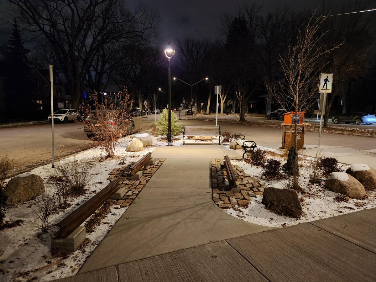 Image showing 14th Street parklet after construction at night with snow on the ground. The parklet feature decorative heritage rails, new concrete walkway, plantings, benches, bike racks, and lighting.